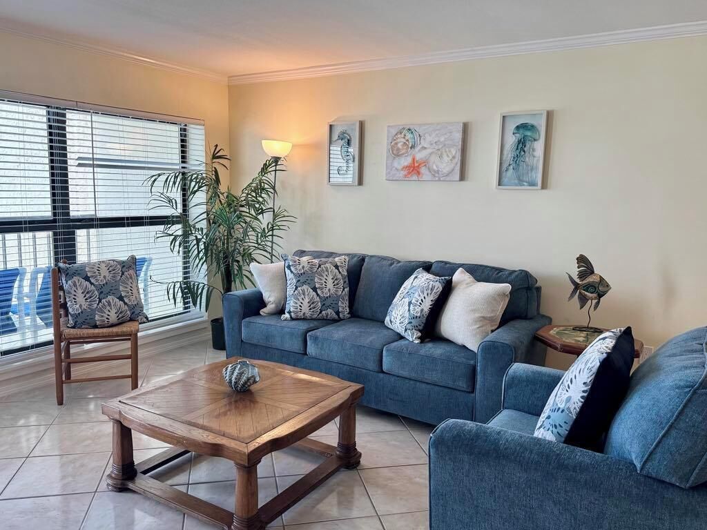 Una sala de estar con un sofá azul, dos sillones, una mesa de centro de madera y una lámpara de pie. Se observan tres cuadros colgados en la pared y una planta en maceta.