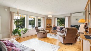 A spacious living room with a sofa, armchairs, a coffee table, and a television. Large windows provide natural light and a view of greenery outside. - Charming 3BR Home with River Views & Sauna (Rovaniemi)