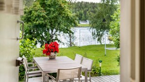 An outdoor dining area with a table and chairs, a vase of red flowers, and a view of a lake and greenery. - Charming 3BR Home with River Views & Sauna (Rovaniemi)