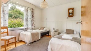 A bedroom with two beds, a wooden chair, a nightstand, a clock, and a window with curtains. - Charming 3BR Home with River Views & Sauna (Rovaniemi)