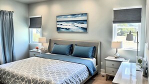 A modern bedroom with a bed, bedside tables, a nightstand, a lamp, a painting of the ocean, and a window with a view.
