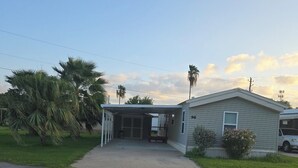Une maison mobile de plain-pied, dotée d’un porche couvert, d’un petit jardin devant et d’un palmier.