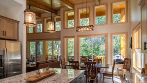 Una cocina amplia con una isla grande, armarios de madera y un refrigerador de acero inoxidable. Varias ventanas ofrecen luz natural y vistas de los árboles circundantes.