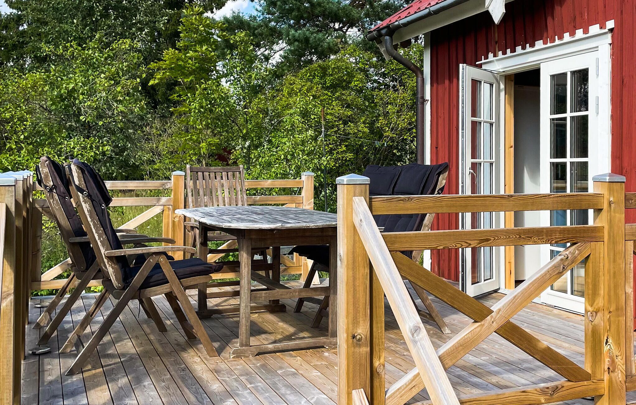 Una terraza de madera al aire libre con una mesa y sillas, un edificio rojo con una puerta blanca y una valla de madera.