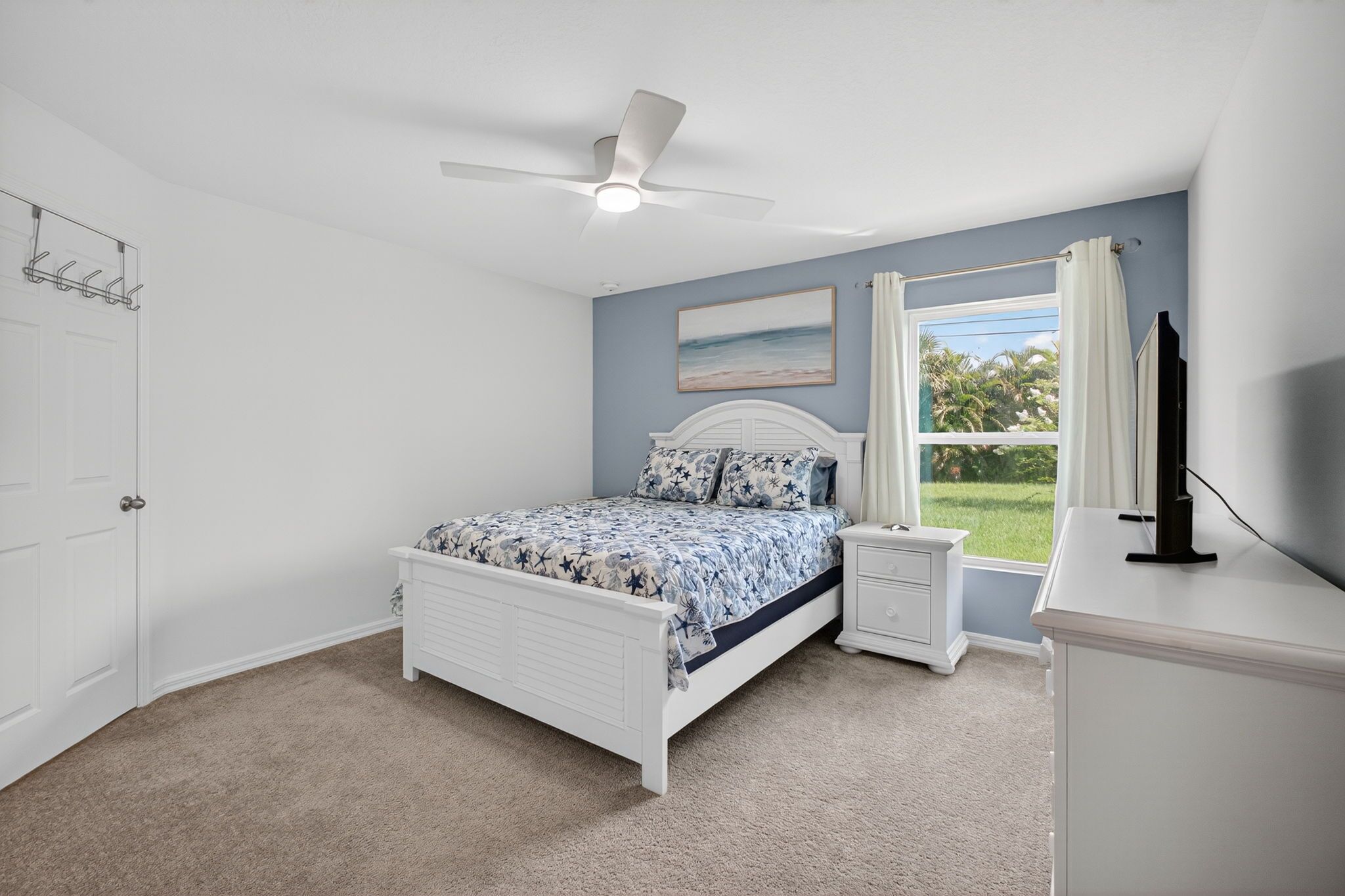 A bedroom with a bed, nightstands, a TV, a ceiling fan, a window with a view, and a door.