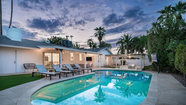 A swimming pool with lounge chairs, a covered patio, and a view of palm trees.