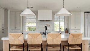 A modern kitchen with white cabinets, a central island with bar stools, pendant lighting, and a view of greenery outside.