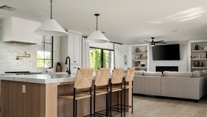 A modern kitchen with a central island, bar stools, a large window, and a living area with a sofa and TV.