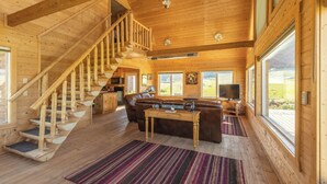 Interior de una cabaña de madera con escalera, sala de estar con sofá, mesa de centro y televisor.
