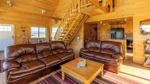 Interior de una cabaña de madera con dos sofás de cuero, una mesa de centro, una escalera, una cocina y vistas a través de una ventana.