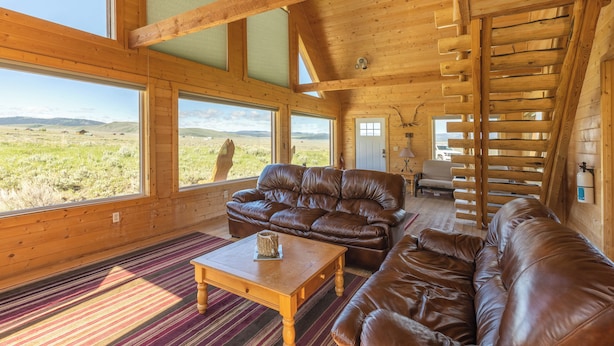 Una habitación amplia con un sofá de cuero grande, una mesa de centro de madera y vistas a un paisaje cubierto de hierba a través de amplios ventanales.