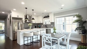 Una cocina moderna con armarios blancos, electrodomésticos de acero inoxidable, una isla central con taburetes de bar y un comedor con alfombra de diseño tipo mármol.