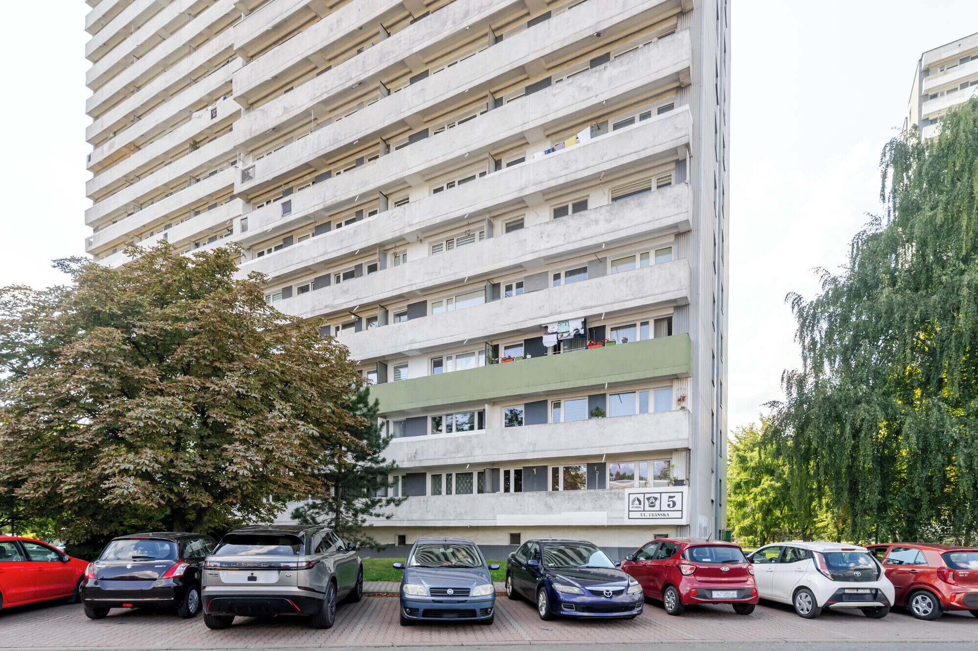 A multi-story residential building with balconies, parked cars, and trees.