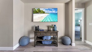 A room with a wooden console table, two round blue poufs, a framed picture of a beach, and a glimpse of a bedroom with a bed and a nightstand.