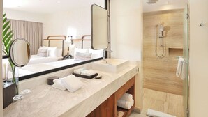 A modern hotel bathroom with a large mirror, two sinks, a shower, and a bed with a headboard.