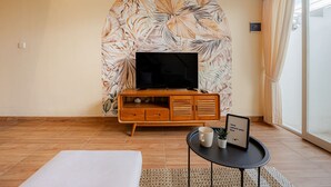 A living room with a flat-screen TV on a wooden cabinet, a small table with a mug and a framed picture, and a large window with curtains. - Cove Lotus Loft Seminyak (Keroboken Kelod)