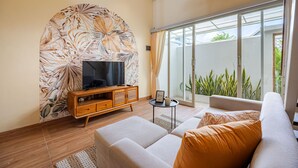 A modern living room with a flat-screen TV, a wooden cabinet, a sofa, and a large sliding glass door leading to a balcony with plants. - Cove Lotus Loft Seminyak (Keroboken Kelod)