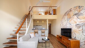 A modern living room with a staircase, a sofa, a TV, and a kitchen in the background. - Cove Lotus Loft Seminyak (Keroboken Kelod)