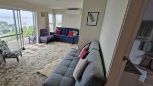 Ein geräumiges Wohnzimmer mit einem großen Fenster und Blick ins Freie, ein graues Sofa mit roten und weißen Kissen, ein blaues Sofa mit roten Kissen, ein kleiner runder Tisch und eine Holzkommode.