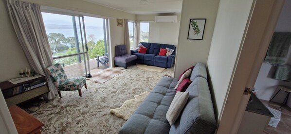 A spacious living room with a large window offering a view of the outdoors, a grey sofa with red and white cushions, a blue sofa with red cushions, a small round table, and a wooden cabinet. - Glorious views and relaxation a plenty (Kawhia)