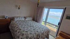 Ein Schlafzimmer mit Bett, Schminktisch, Nachttisch, Fenster mit Meerblick und einem Bild an der Wand.