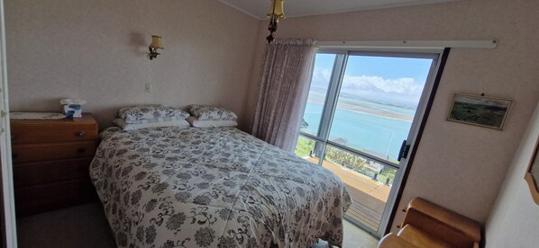 A bedroom with a bed, a dresser, a nightstand, a window with a view of the sea, and a picture on the wall. - Glorious views and relaxation a plenty (Kawhia)