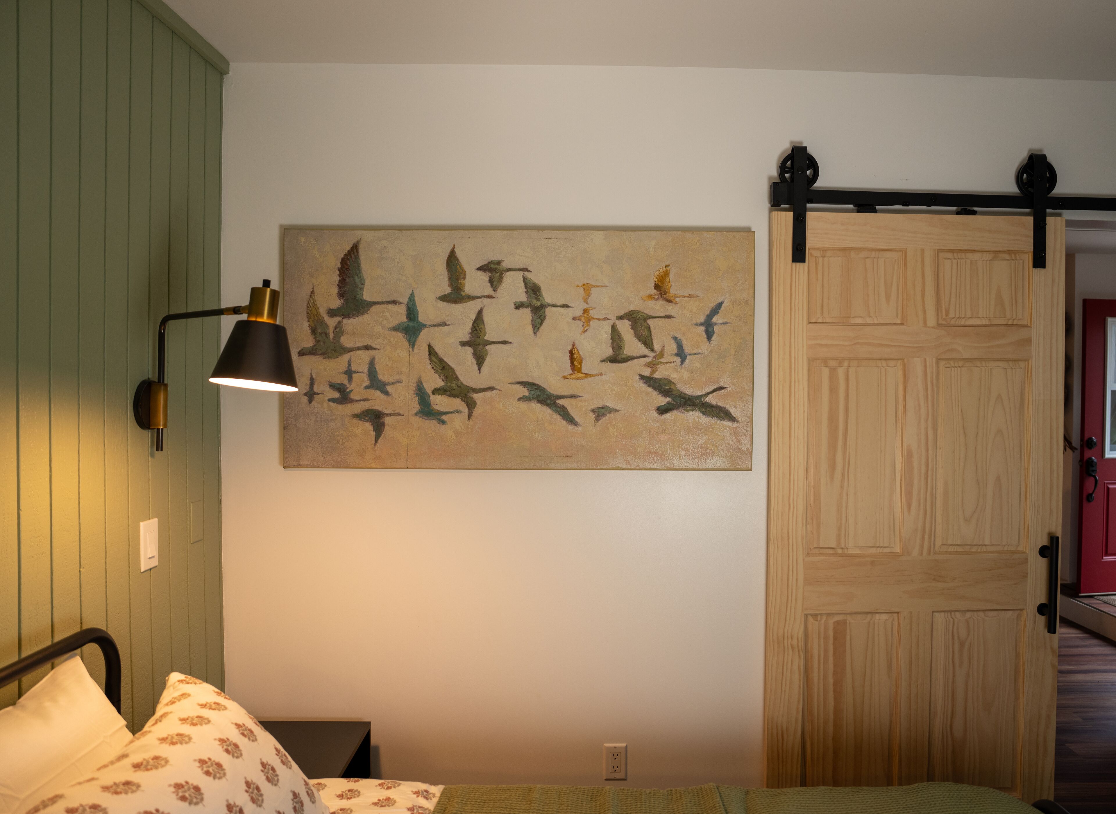 A bedroom with a bed, a wall-mounted lamp, a wooden door, and a painting of birds.