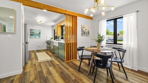 A modern kitchen with wooden flooring, a dining area with a round table and chairs, and a view of the outside through a large window.