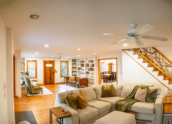 A spacious living room with a large sectional sofa, a wooden staircase, and a bookshelf filled with books.