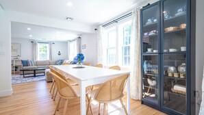 A dining area with a white table and wicker chairs, a glass cabinet with dishes, and a living room with a sofa and coffee table in the background.