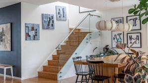 Um interior moderno com escada de madeira, área de jantar com mesa e cadeiras de madeira, e diversas obras de arte emolduradas nas paredes.