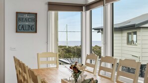 Un coin repas avec une table et des chaises en bois, un panneau encadré accroché au mur et une vue sur la plage, visible à travers de grandes fenêtres.
