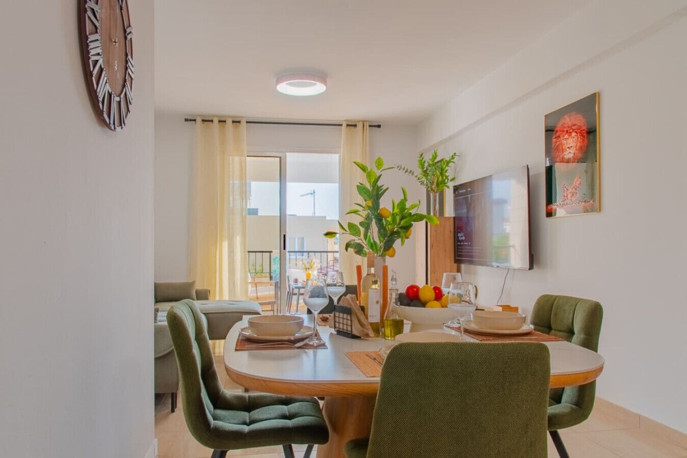 A dining area with a round table, green upholstered chairs, a vase with plants, and a bowl of fruit.