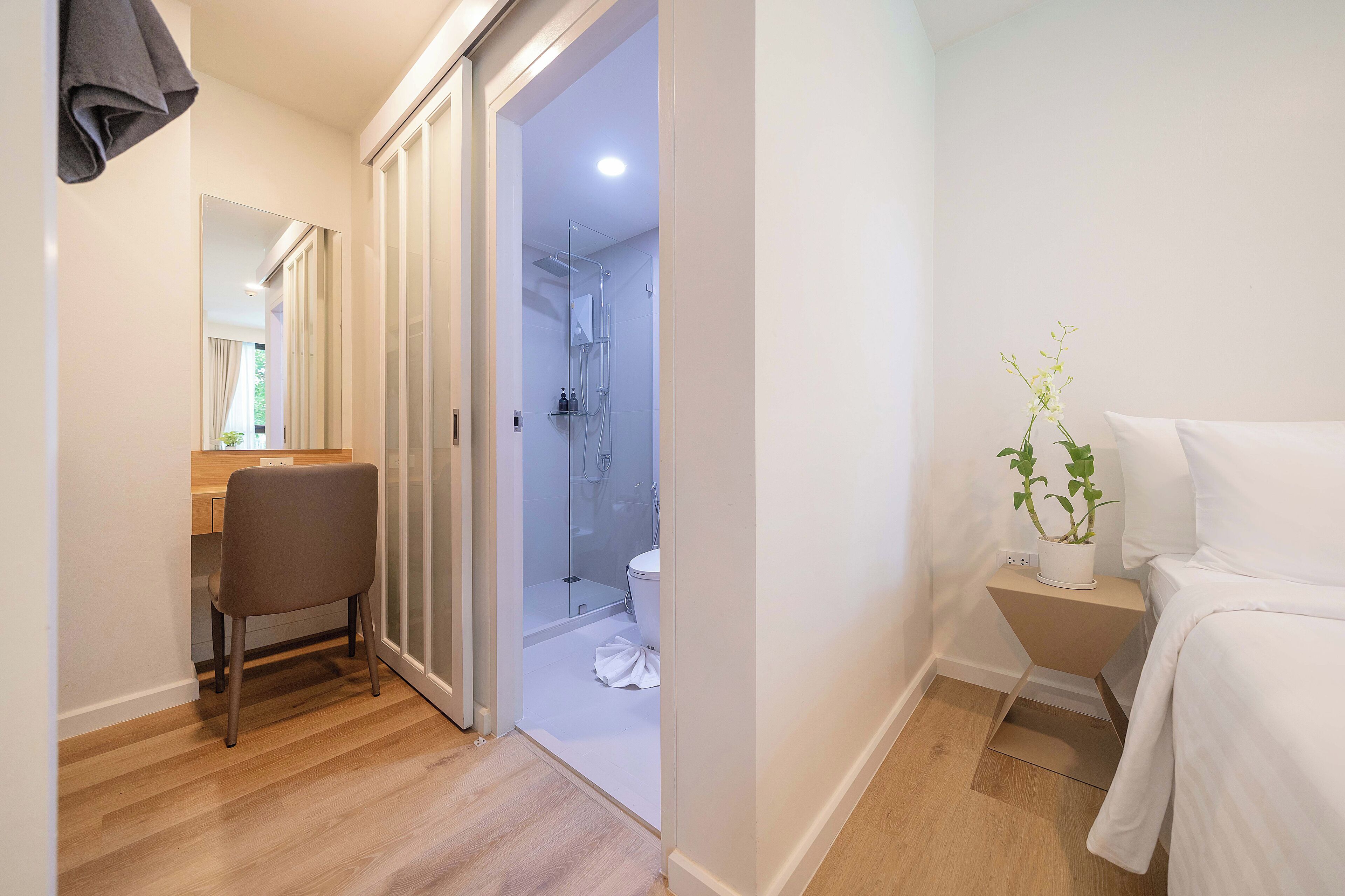 A hotel room with a wooden floor, a bed, a bedside table with a plant, a bathroom with a shower, and a desk with a chair.