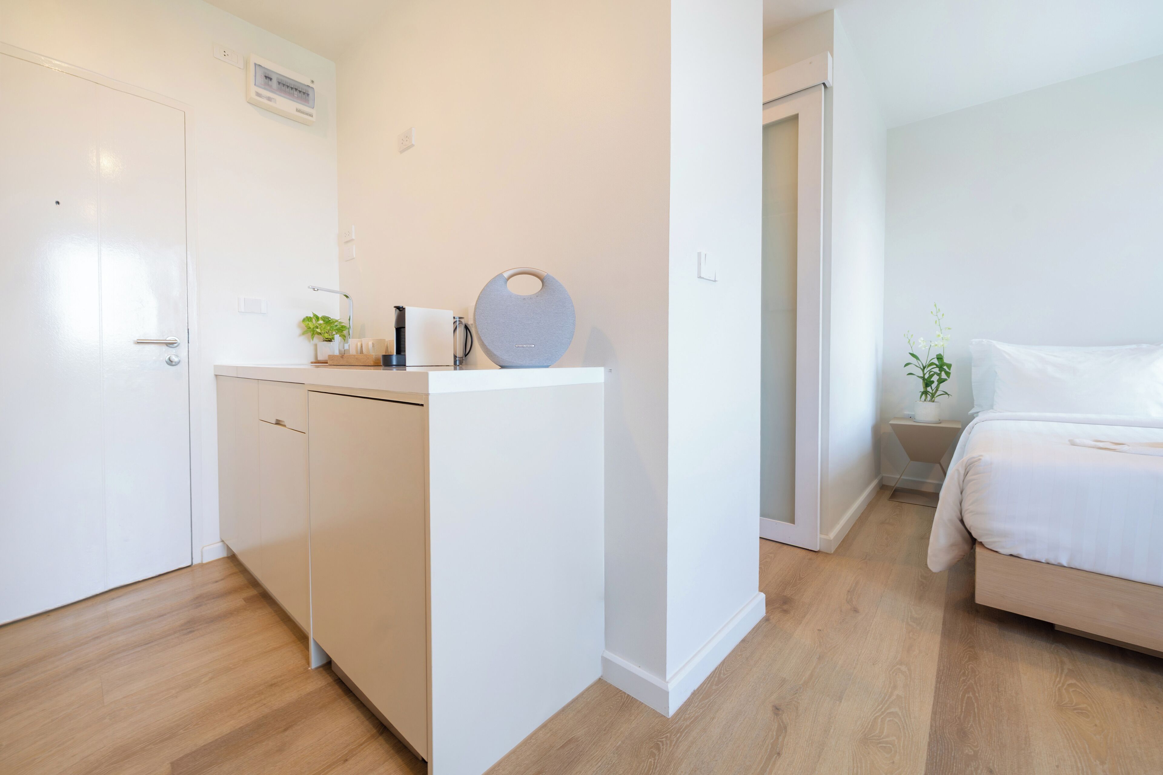 A compact hotel room with a kitchenette, a bed, and a small bedside table with a plant.