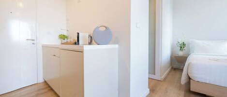 A compact hotel room with a kitchenette, a bed, and a small bedside table with a plant.