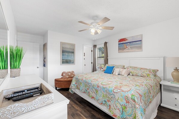 A bedroom with a bed, a ceiling fan, a nightstand, a lamp, a painting, and a window with blinds. - Sea Grove 307 - Walk to Surfside Beach! (Surfside Beach)