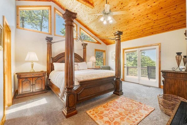 A four-poster bed with a wooden headboard, a ceiling fan, and a large window with a view of trees.