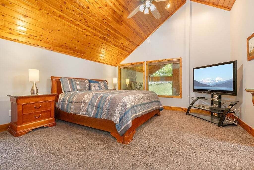 A bedroom with a wooden ceiling, a bed with patterned bedding, a nightstand, a TV on a stand, and a window with a view of mountains.