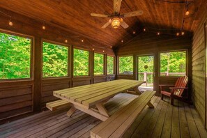 A wooden deck with a picnic table and chairs, large windows with a forest view, and a ceiling fan.