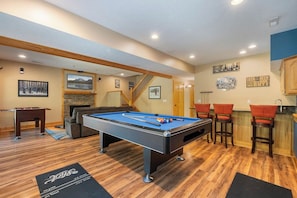 Una mesa de billar en una habitación con suelo de madera, un sofá, una escalera y una zona de bar.