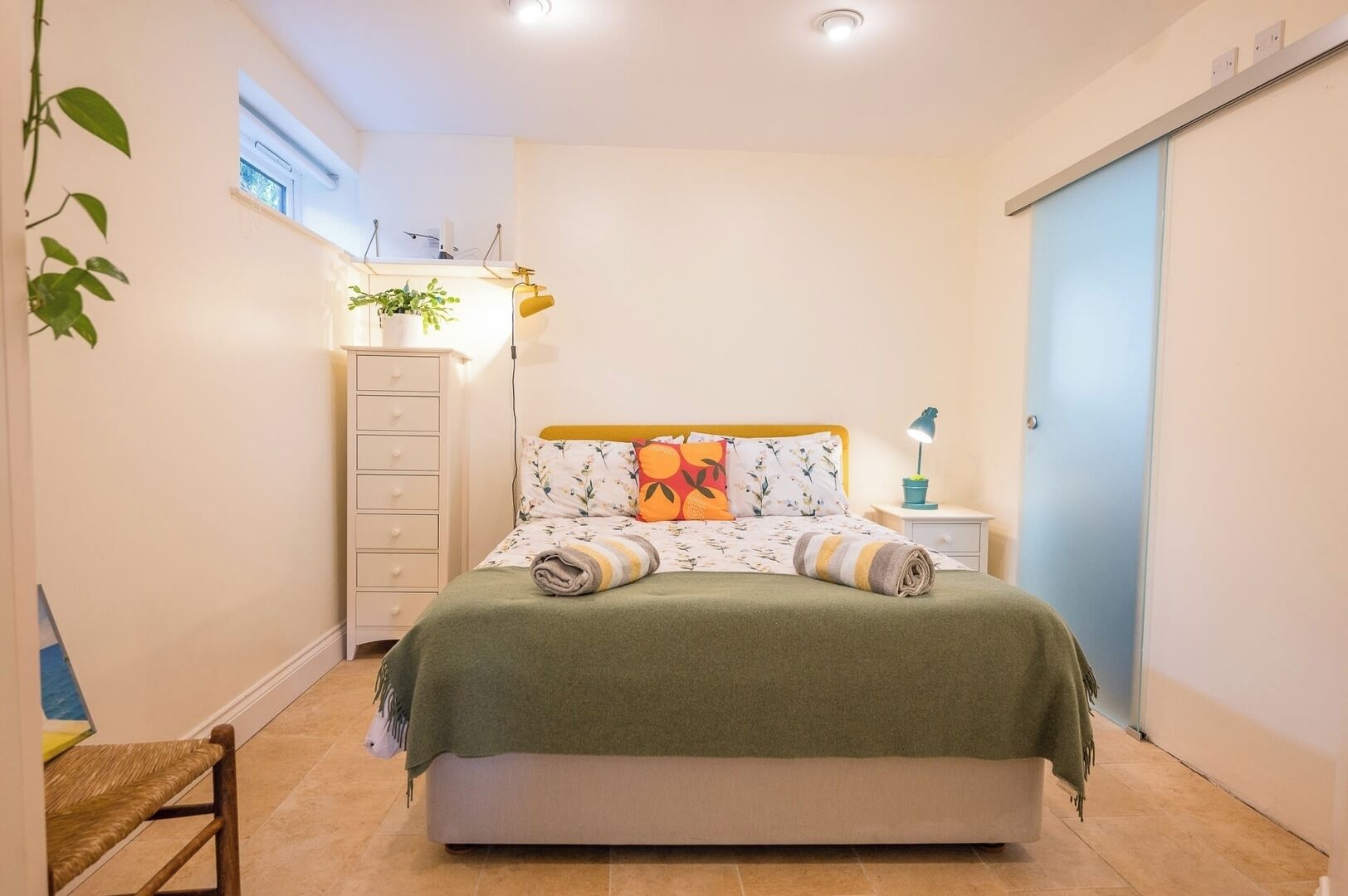 Une chambre à coucher avec un lit, une table de chevet, une commode, une fenêtre et une plante.