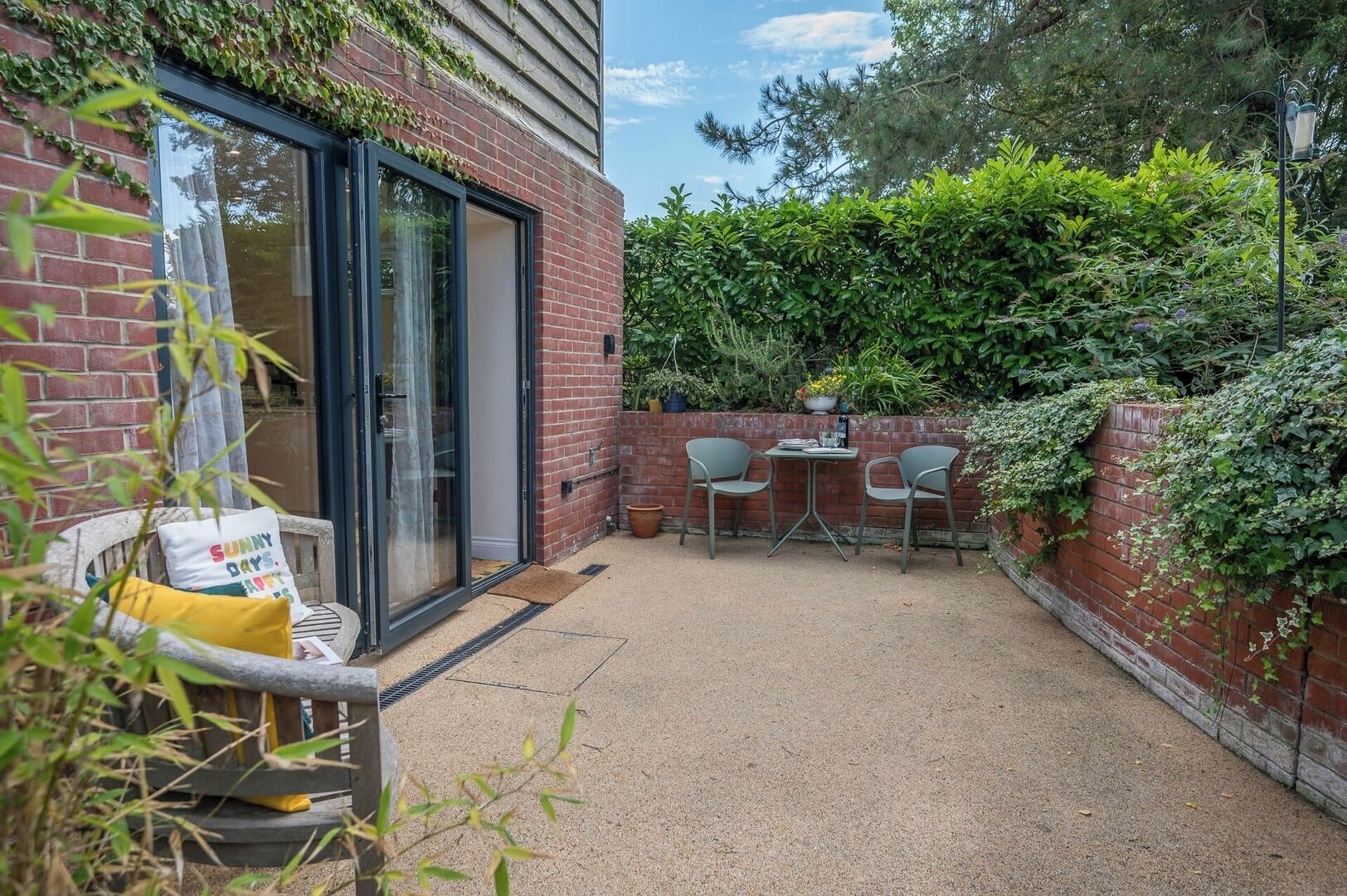 Eine Terrasse mit Tisch und Stühlen, eine Glastür und eine Ziegelwand mit Grünpflanzen.