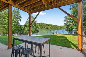 A covered patio with a table and chairs, overlooking a lake and forested area. - Dockside of the Moon | Lakefront Luxury, Views & Private Dock (Nebo)