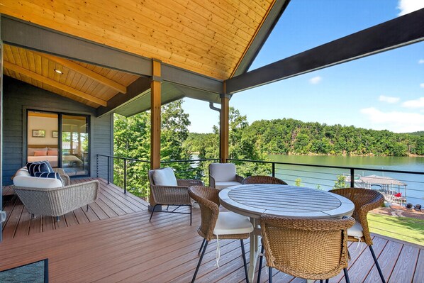 A wooden deck with wicker furniture and a round table, overlooking a lake and forested hills. - Dockside of the Moon | Lakefront Luxury, Views & Private Dock (Nebo)