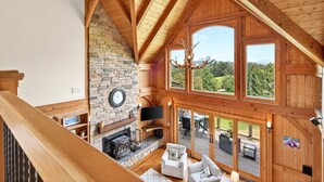 A spacious living area with a stone fireplace, a television, and a large window offering a view of the outdoors. - Windy Path Farm | Animals, Hot Tub, Game Room + Breathtaking VIEWS! (Fairview)