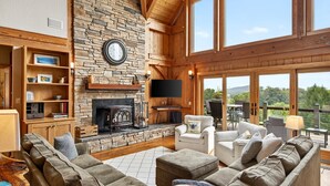 A spacious living room with a stone fireplace, wooden beams, and large windows overlooking a scenic landscape. - Windy Path Farm | Animals, Hot Tub, Game Room + Breathtaking VIEWS! (Fairview)