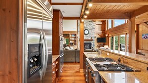 A modern kitchen with stainless steel appliances, a central island with a stove, wooden cabinetry, and a stone accent wall. - Windy Path Farm | Animals, Hot Tub, Game Room + Breathtaking VIEWS! (Fairview)
