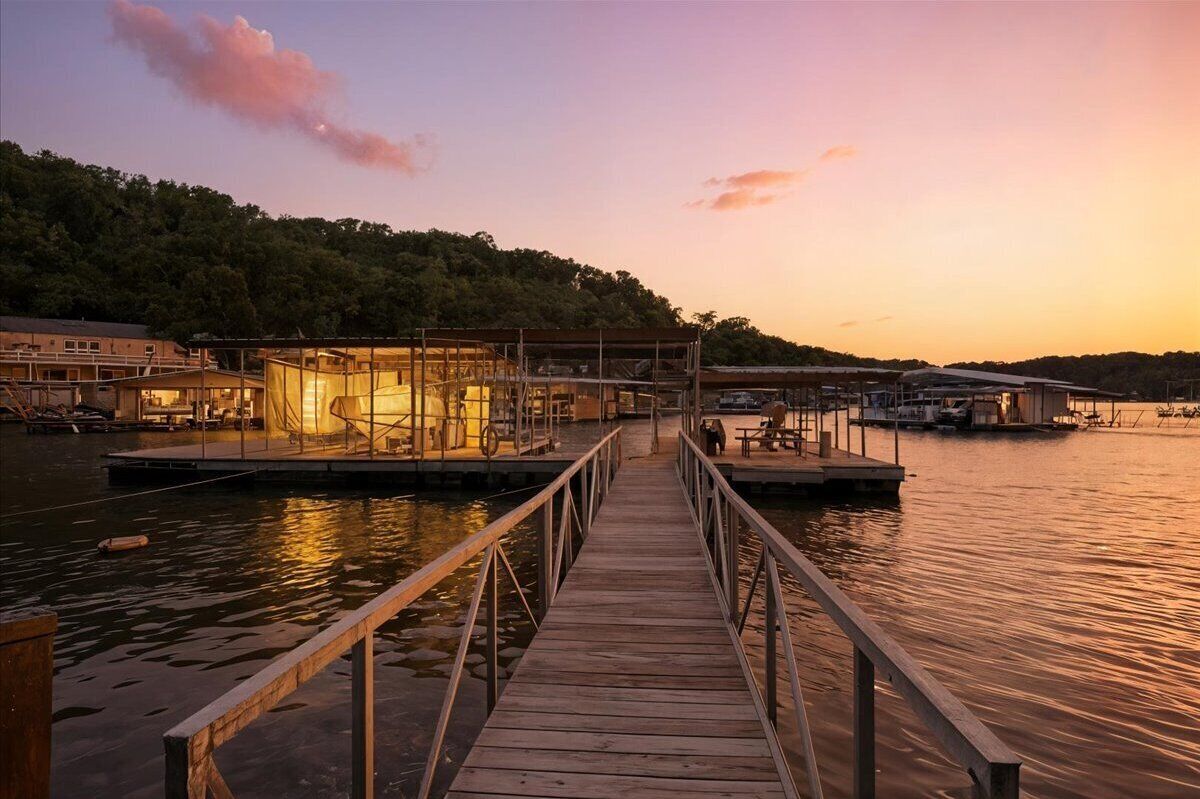 Un muelle de madera que conduce a un edificio con paredes de vidrio y un embarcadero que se extiende sobre aguas tranquilas, rodeado de árboles y otras construcciones.