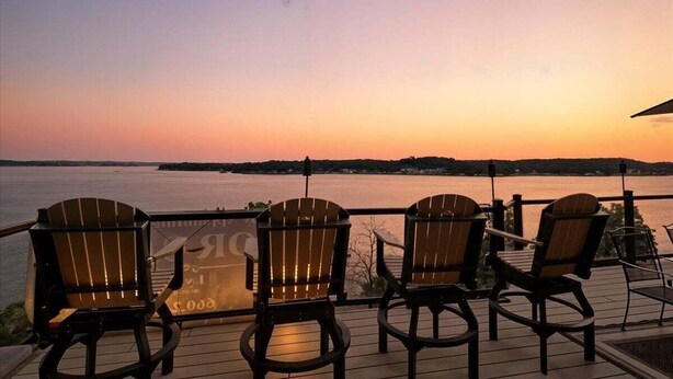 A deck with Adirondack chairs overlooking a body of water during sunset.